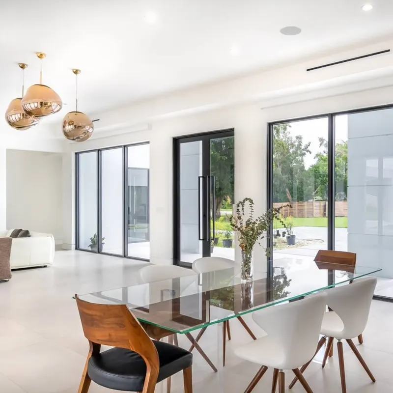Bright dining room with a glass table and white chairs beside floor-to-ceiling dark-framed glass doors looking onto a lawn