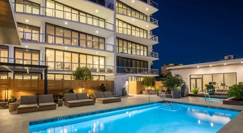 Multi-story residential building with rows of lit windows and glass balcony doors above a pool deck at dusk