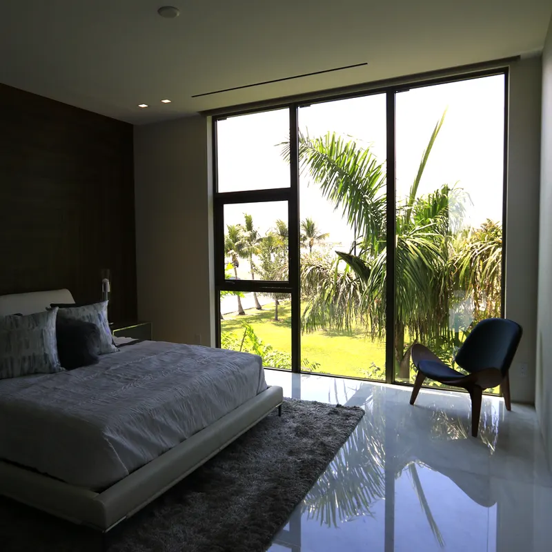 Modern bedroom with a low bed and large dark-framed glass panels overlooking palms and a lawn