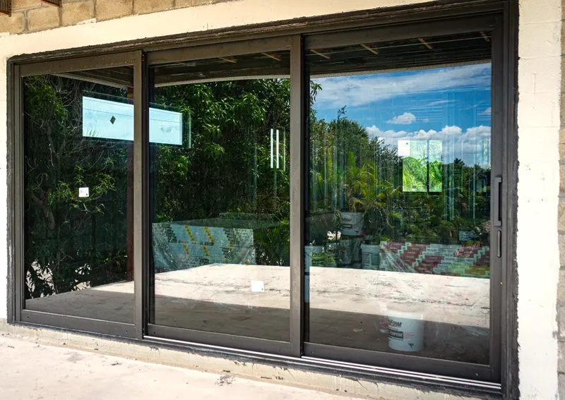 Newly installed dark-framed multi-panel sliding glass door in a stucco wall, with protective film and labels still on the glass