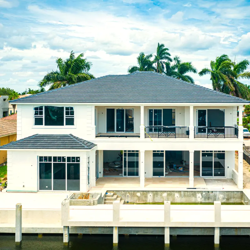 Two-story white waterfront house with many dark-framed windows and doors facing a dock, palm trees behind