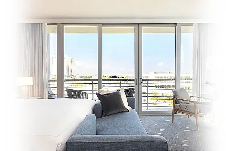 Living room with a gray sofa and armchair facing floor-to-ceiling sliding glass doors that open to a balcony with a city view