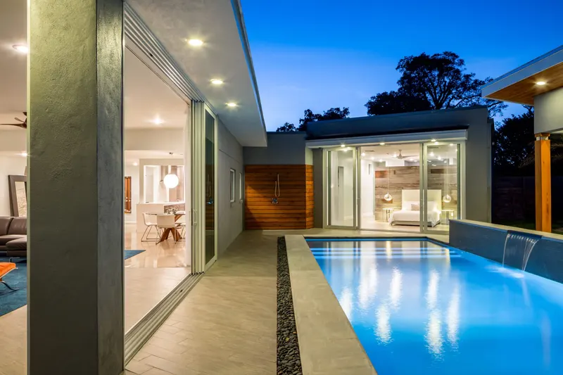 Modern home at dusk with open sliding glass doors beside a lit lap pool